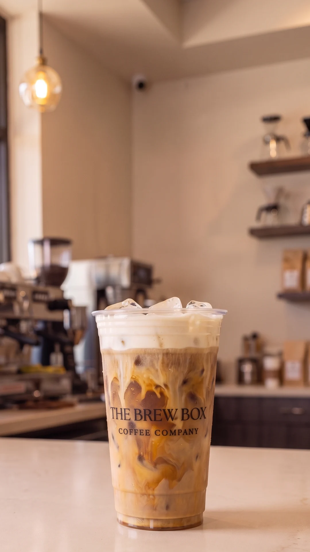 A tall clear Brew Box takeaway cup of iced shaken espresso with oat milk, caramel marbling and a thick vanilla sweet cream cold foam cap, standing on the service counter in front of a café espresso machine. Concept photography created for this page, not a photograph of the Brew Box menu.